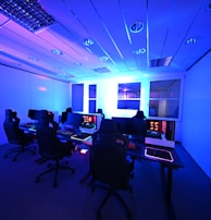 A modern gaming setup with multiple high-backed gaming chairs and monitors arranged in a room with a cool blue lighting. The computers have illuminated keyboards and glowing screens, creating a high-tech ambiance.