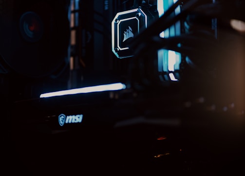 A close-up view of a computer gaming rig illuminated with blue LED lights. The interior shows components like cooling fans at the top, a glowing logo on a central component, and an MSI branded part at the bottom. The scene is dark, emphasizing the glow of the lights.