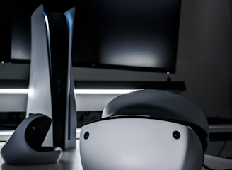A sleek virtual reality headset sits on a reflective surface beside a gaming console and its controller. The background features a large dark monitor screen, creating a modern and futuristic setting. The lighting is subdued, highlighting the glossy surfaces of the electronic devices.