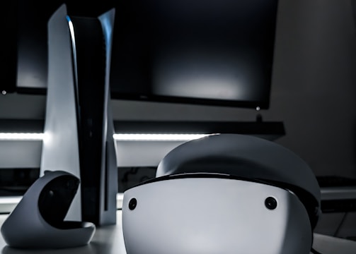 A sleek virtual reality headset sits on a reflective surface beside a gaming console and its controller. The background features a large dark monitor screen, creating a modern and futuristic setting. The lighting is subdued, highlighting the glossy surfaces of the electronic devices.